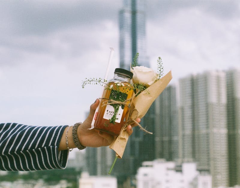Hand holding a jar of iced tea with a straw and a bouquet featuring a white rose and greenery, set against a city skyline, symbolizing thoughtful gift ideas for tech-savvy adults.