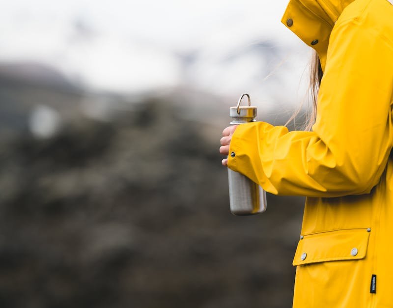 Person in a yellow rain jacket holding a stainless steel water bottle, set against a blurred outdoor background, reflecting an active lifestyle and outdoor adventures.