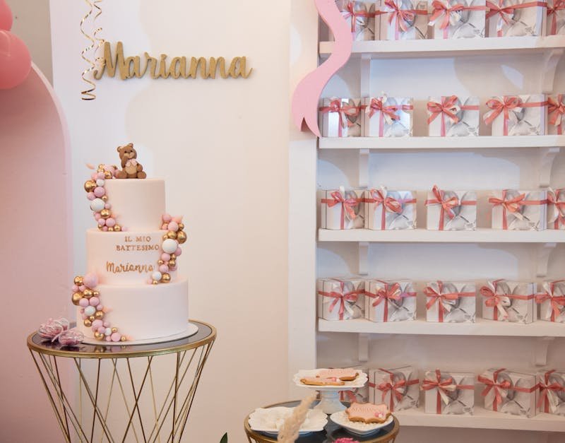 Cake decorated for a baptism celebration with a teddy bear, pink and gold accents, and personalized name "Marianna," surrounded by gift boxes on a display shelf.