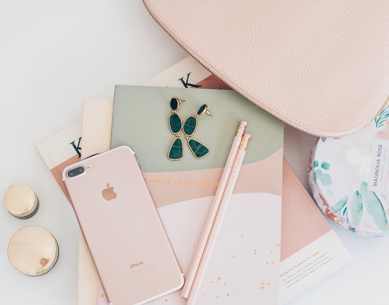 Pink smartphone, gold and green earrings, two pink pencils, and a textured pouch on colorful notebooks, representing modern lifestyle and organization for tech-savvy adults.