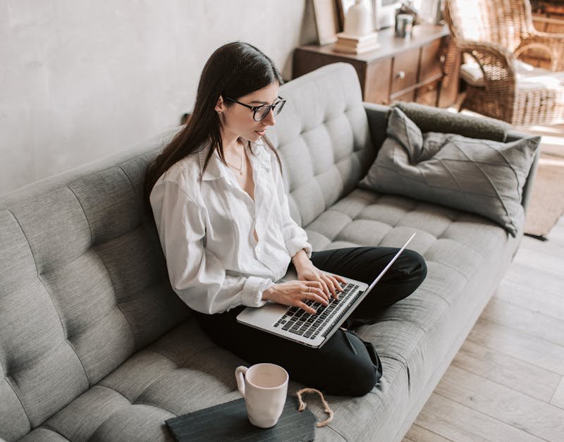 Woman in casual attire working on a laptop while sitting on a gray sofa, with a coffee mug and a notepad nearby, illustrating a comfortable home office environment.