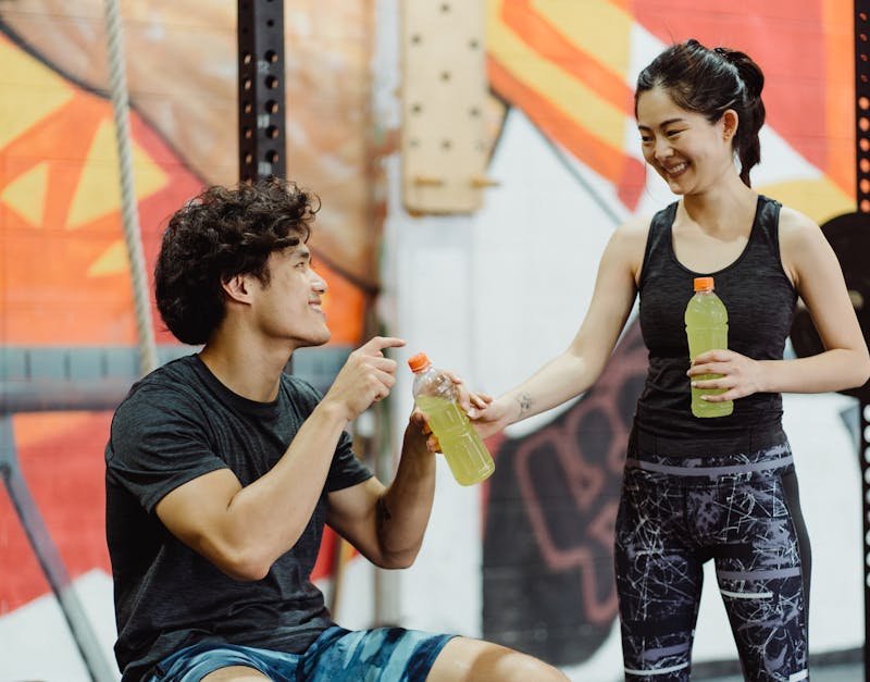 Young man and woman in athletic wear sharing sports drinks in a gym setting, emphasizing fitness and hydration, relevant to high-tech gadgets and thoughtful gift ideas for women.