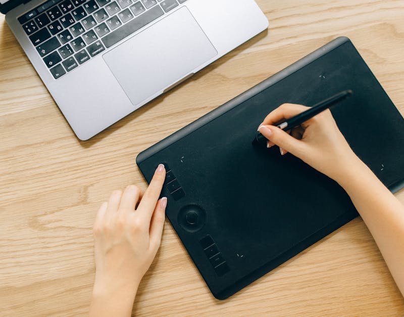 Person using a graphics tablet with stylus beside a laptop on a wooden desk, illustrating digital art creation and smart home technology integration.
