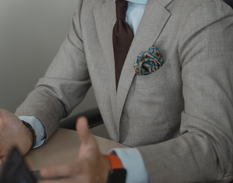 Man in a gray suit with a brown tie and colorful pocket square, gesturing while discussing men's watch styles and trends.