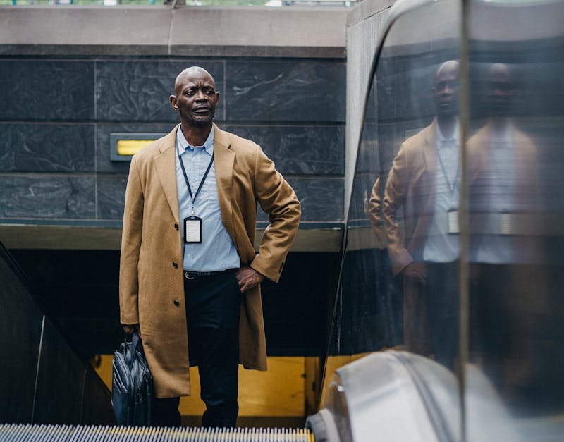 Man in a tan coat standing at the top of an escalator, holding a briefcase, representing a modern professional lifestyle in an urban setting.