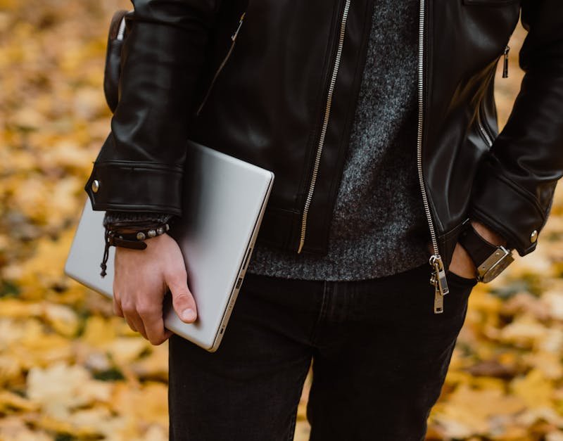 Man in a black leather jacket holding a laptop, surrounded by autumn leaves, representing modern tech and lifestyle in gift ideas for men.