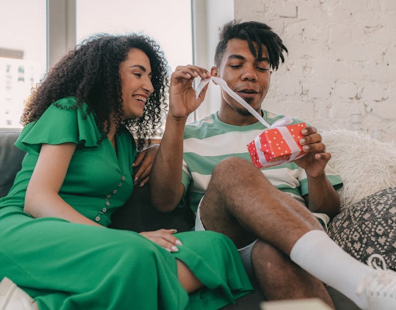 Couple joyfully exchanging gifts, man unwrapping a colorful present while seated on a couch, highlighting thoughtful gift ideas for men.