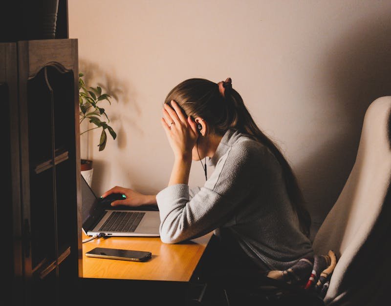 Person sitting at a desk with laptop, covering face in frustration, wearing headphones, in a home office setting, with a plant and smartphone visible, reflecting the challenges of using high-tech gadgets.