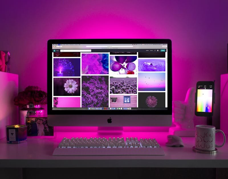 Desktop computer with a vibrant purple-themed display showcasing various floral and abstract images, illuminated by pink backlighting, alongside a smartphone and a decorative mug, reflecting a creative workspace ideal for content creation and photography enthusiasts.