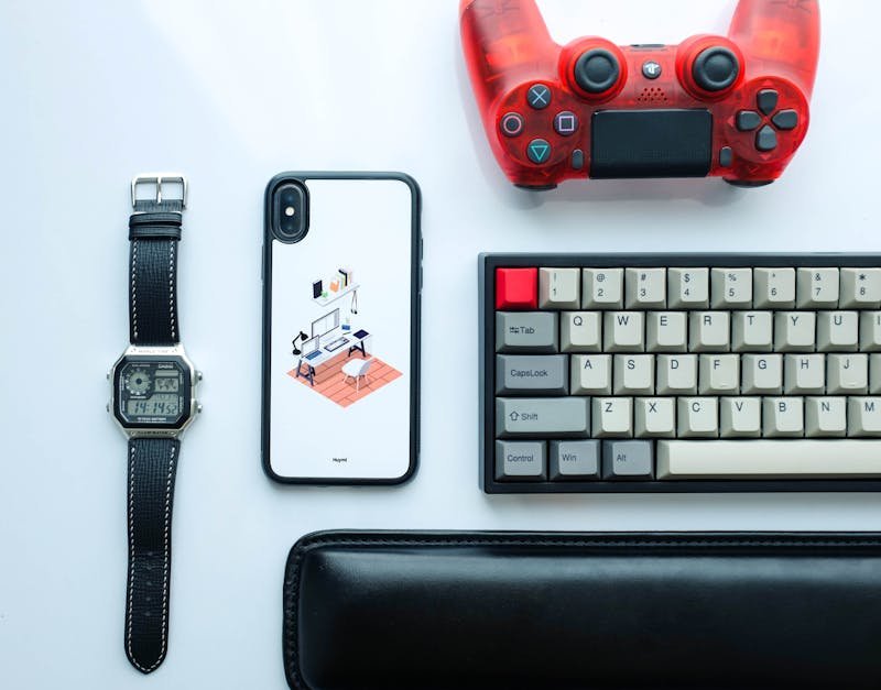 Wireless gaming accessories including a mechanical keyboard, gaming controller, smartphone case with a desk illustration, and a digital watch on a white background.