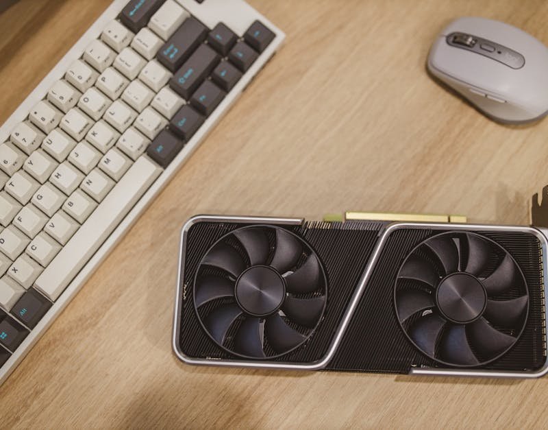 Gaming PC components including a high-performance graphics card with dual fans, a mechanical keyboard, and a computer mouse on a wooden desk.