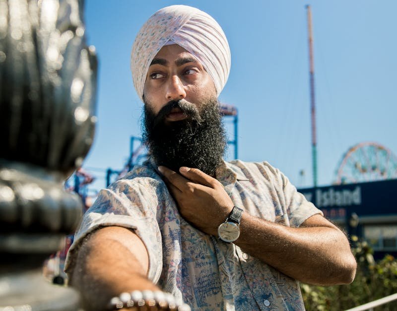 Man wearing a turban and floral shirt, showcasing a stylish wristwatch, with an amusement park in the background.