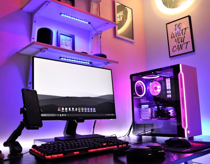 Gaming setup with a desktop monitor displaying a landscape, mechanical keyboard with red backlighting, smartphone on a stand, and a custom PC case featuring RGB lighting.