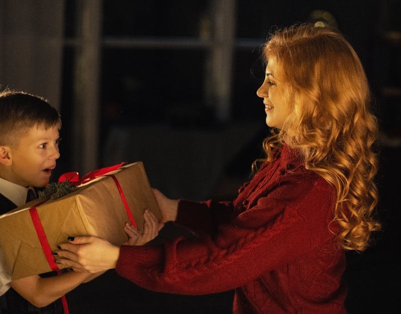 Woman in a red sweater handing a wrapped gift to a boy with a surprised expression, symbolizing joy and celebration.