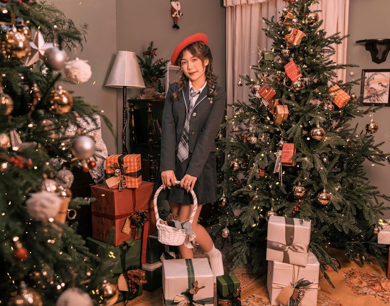 Young woman in a stylish outfit holding a basket, surrounded by Christmas trees and wrapped gifts, creating a festive holiday atmosphere.