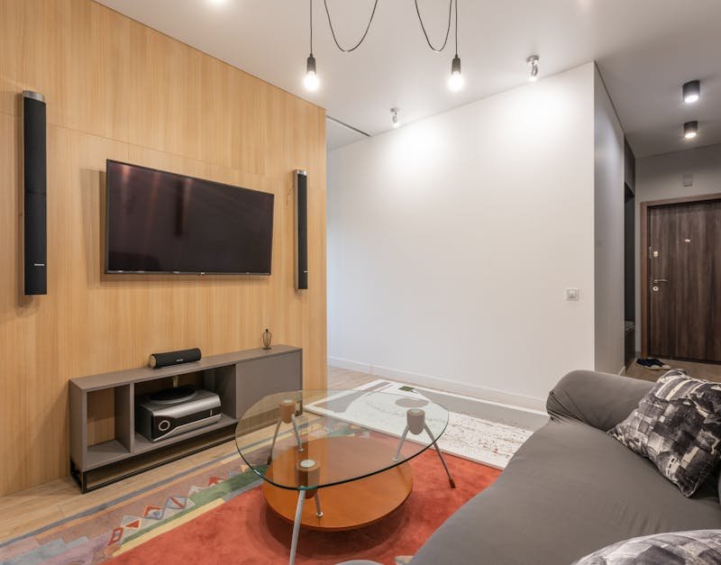 Interior of contemporary light apartment with couch with pillows near table on carpet and cabinet with TV on wall with speakers near lamps on ceiling