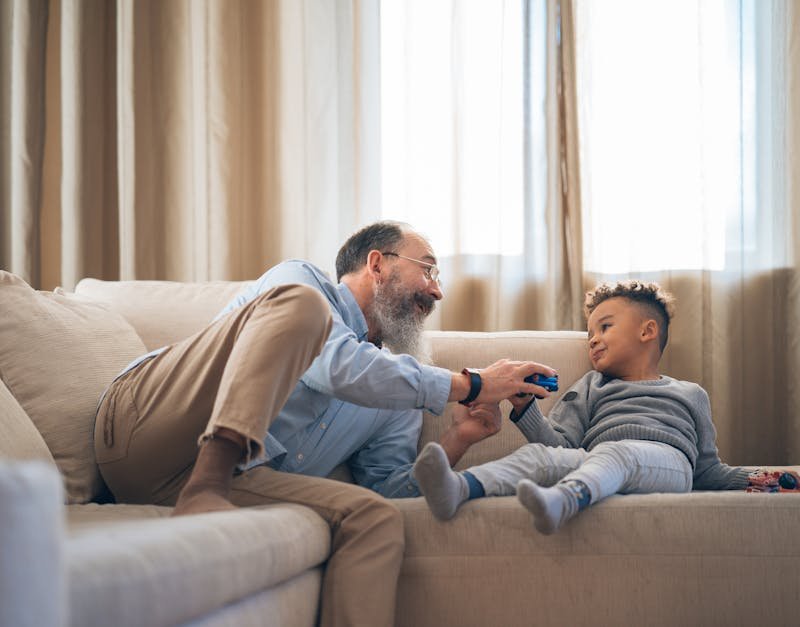 A heartwarming moment between grandfather and grandson laughing and playing on a cozy sofa indoors.