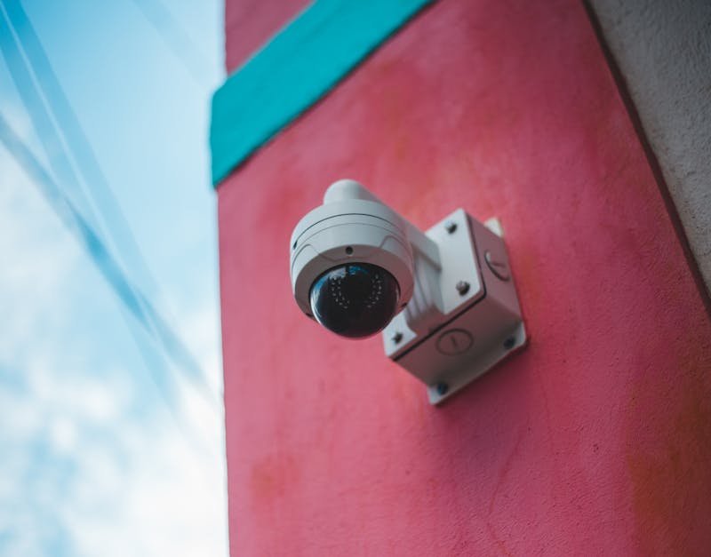 Security camera mounted on a vibrant pink wall, capturing surveillance footage in an urban environment.