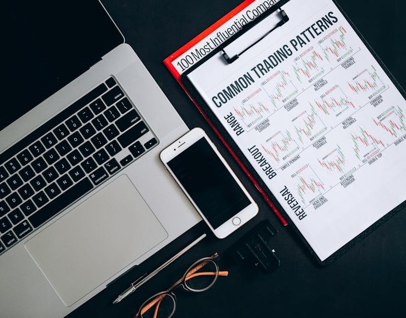 Close-up of office tools including a laptop, smartphone, and trading patterns document on a dark surface.