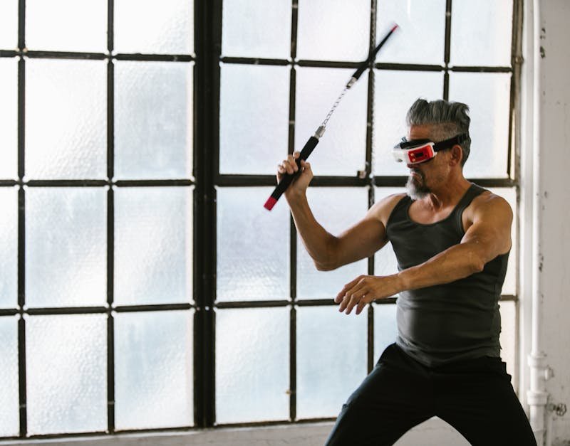 Adult man using VR headset and nunchaku for martial arts training indoors, exploring futuristic technology.