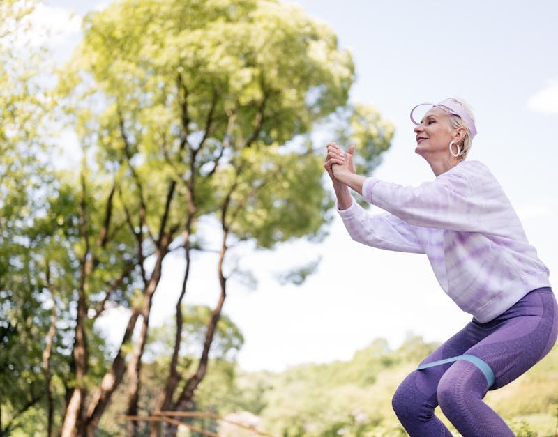 Woman exercising outdoors, performing a squat with resistance band, surrounded by trees, showcasing fitness and wellness.