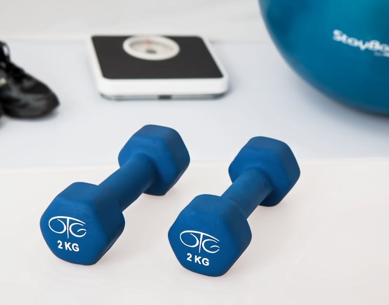 Blue 2 kg dumbbells on a white surface, with a digital scale and exercise ball in the background, representing fitness and home workout equipment for smart home integration.
