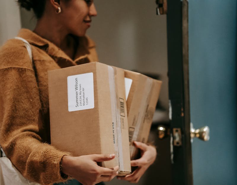Crop ethnic female walking into open door of apartment with carton boxes with goods from delivery
